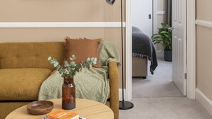 A living room with a beige wall, a framed painting, a sofa, a coffee table with books and a vase, and a door leading to another room. - SACO Bristol- West India House (Bristol)