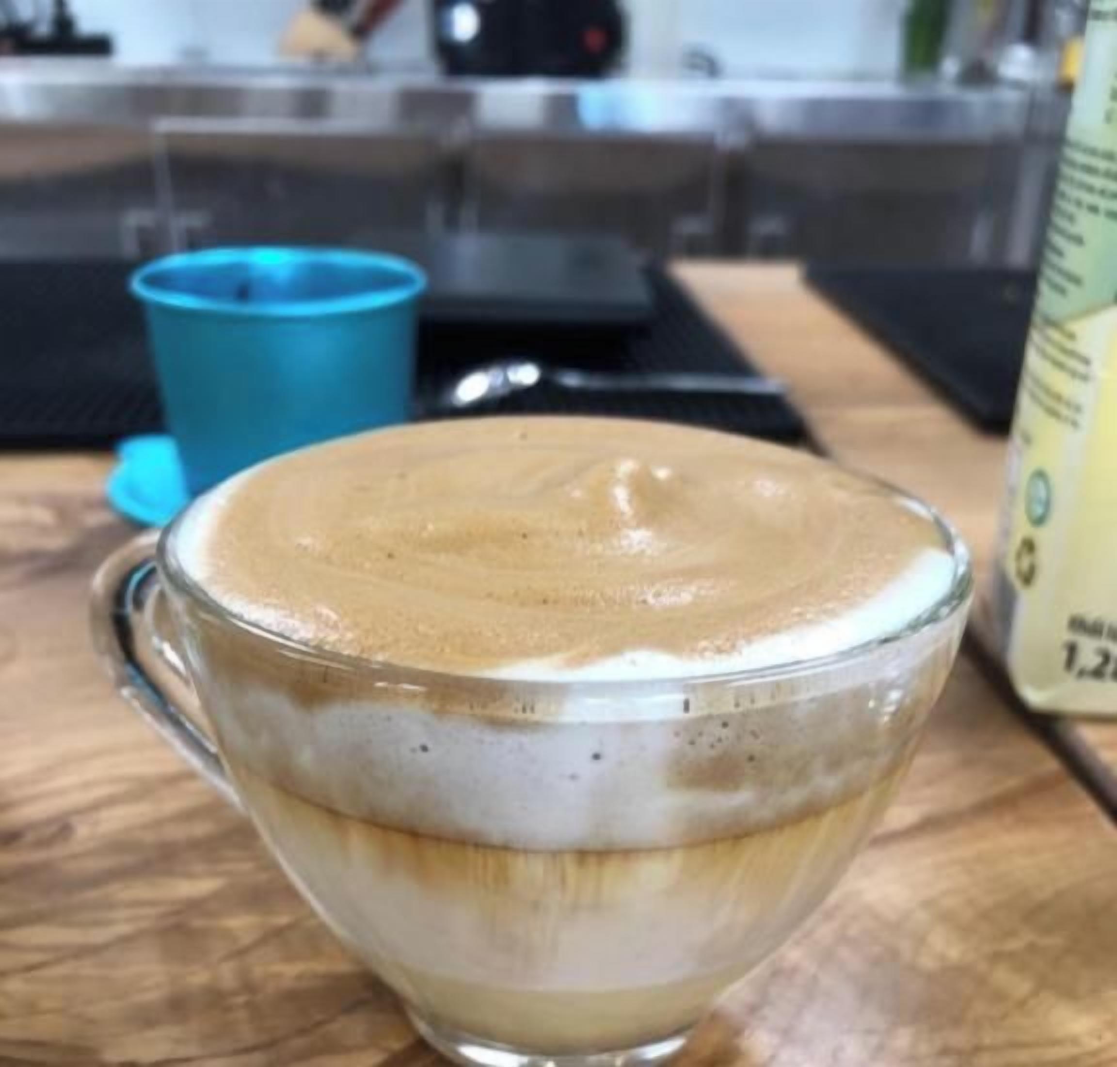 A glass cup of coffee on a wooden surface with a blue cup and a menu in the background.