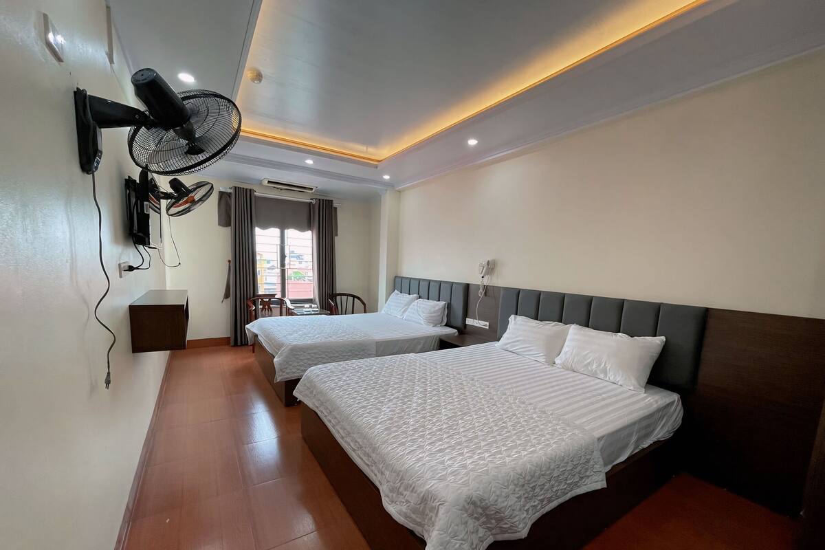 A hotel room with two beds, a wall-mounted fan, an air conditioning unit, and a window with curtains.