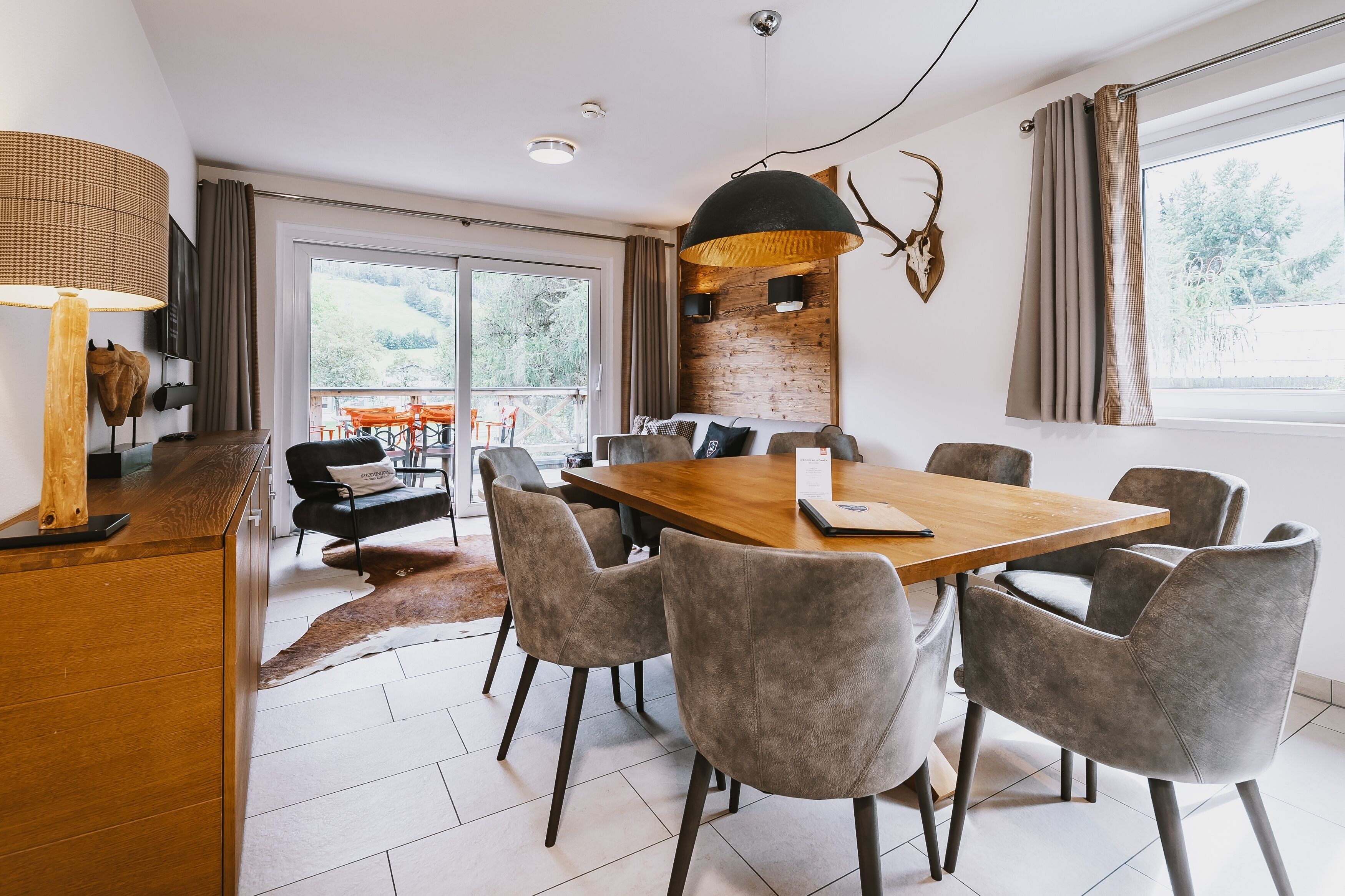 Un espace de repas moderne avec une table en bois et des chaises à revêtement gris, un grand luminaire suspendu et une vue sur l’extérieur grâce à une porte-fenêtre coulissante.
