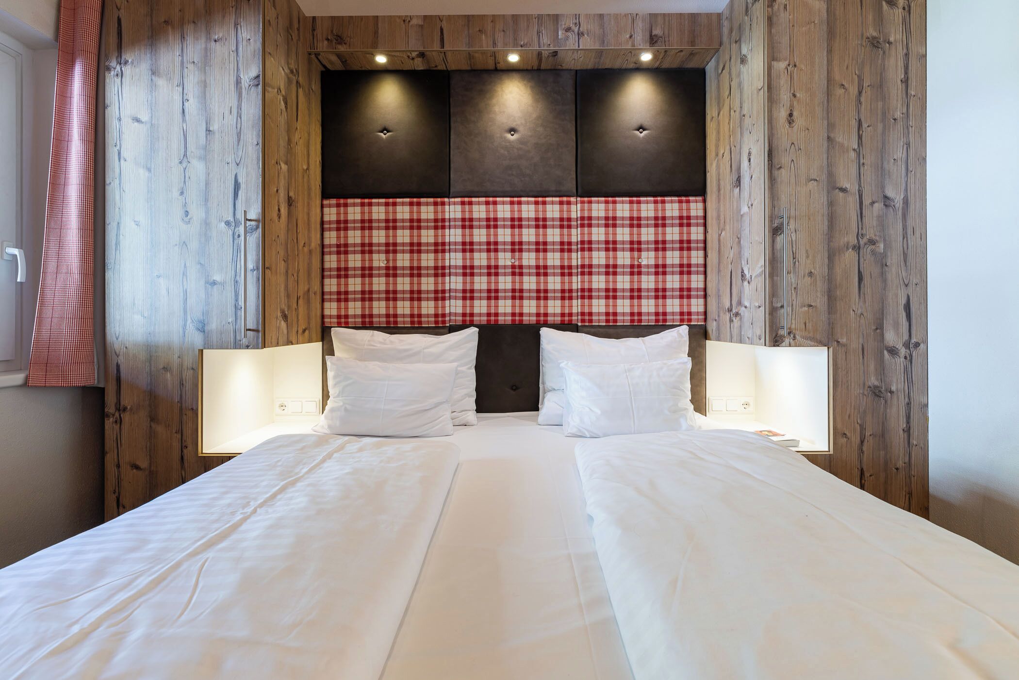 A hotel room with a large bed, wooden panel walls, a red plaid headboard, and two bedside lamps.