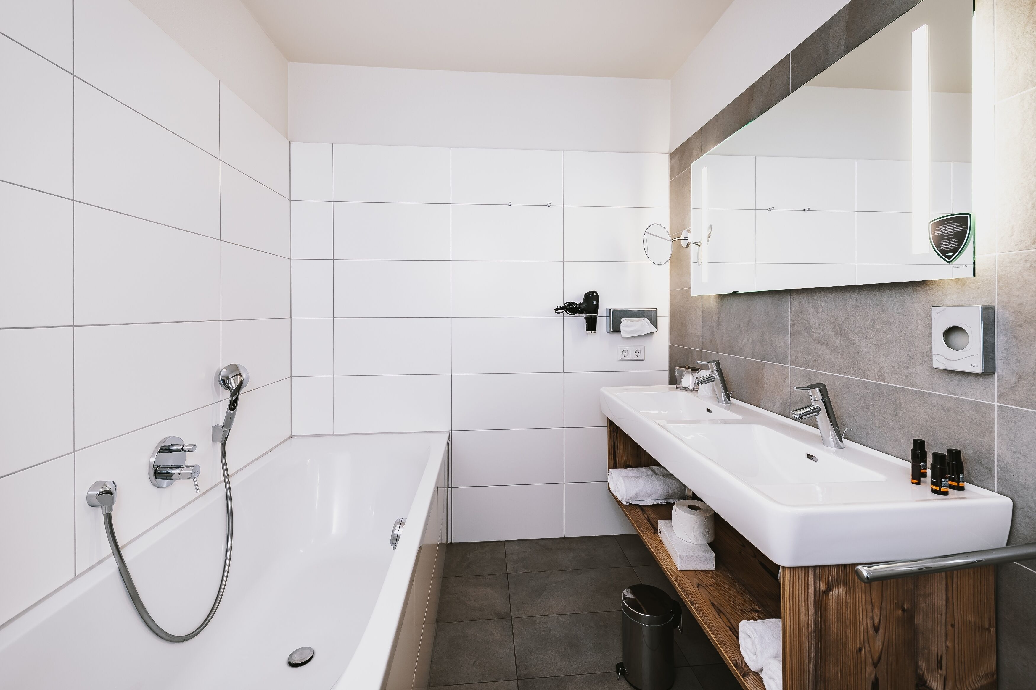 Une salle de bain moderne avec une baignoire, deux lavabos, un miroir et une étagère en bois où sont posées des serviettes.