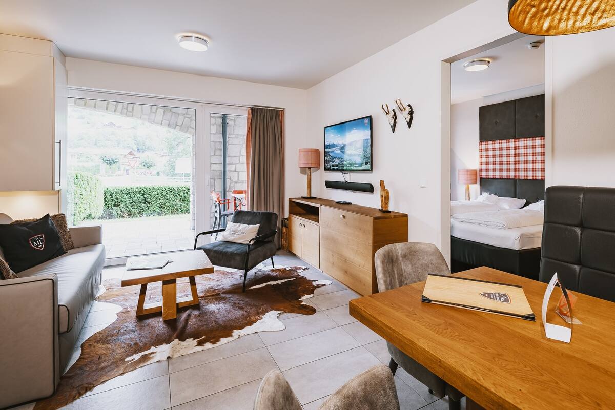 A modern hotel room with a sofa, dining table, and a large window offering a view of the outdoors.