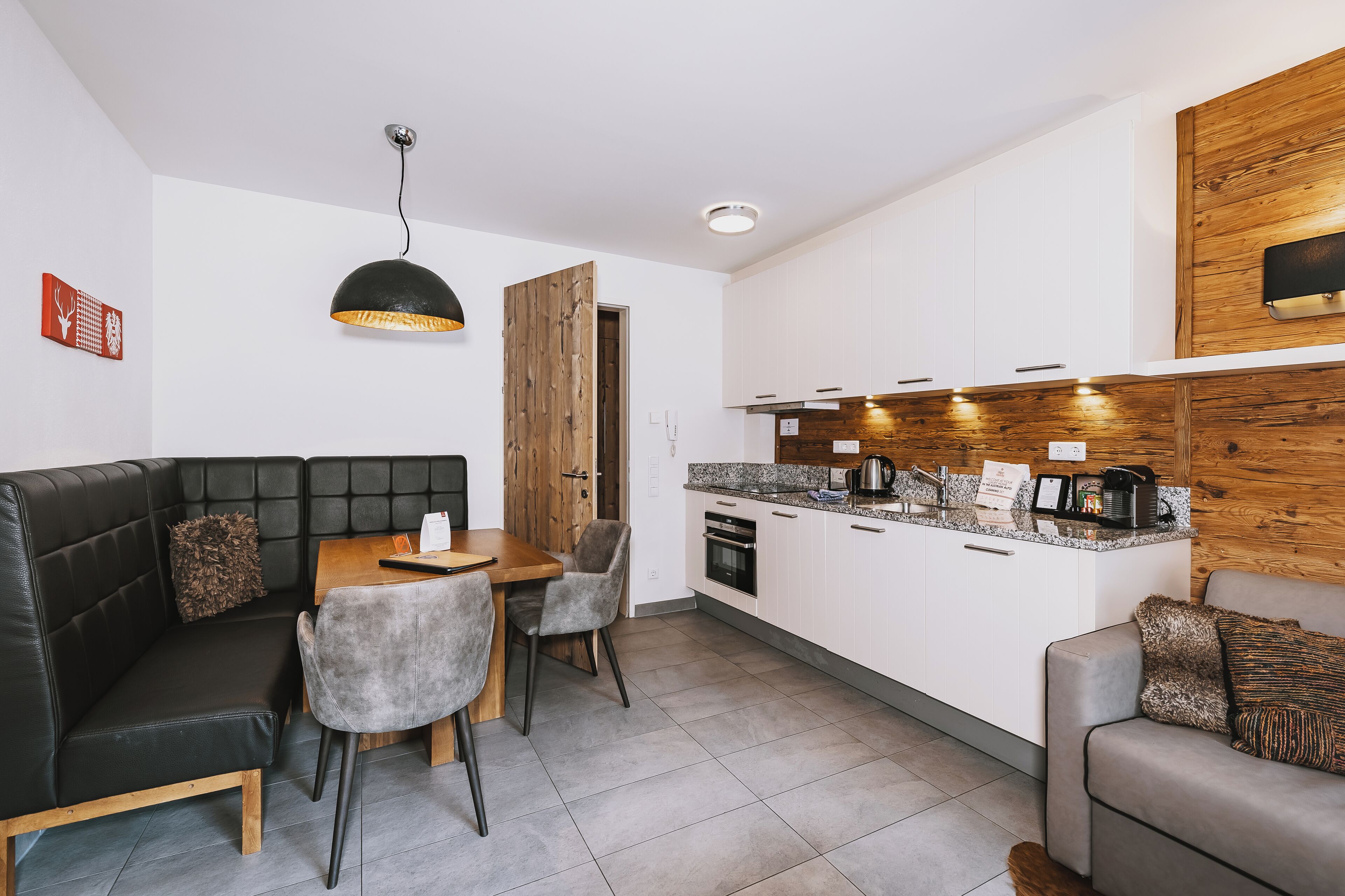 A modern kitchen with white cabinets, a wooden accent wall, a dining area with a black leather sofa, and a grey couch.