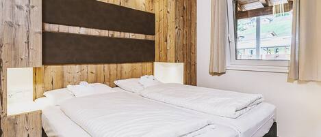 A room with two beds, wooden paneling on the walls, a window with curtains, and a view of the outside.