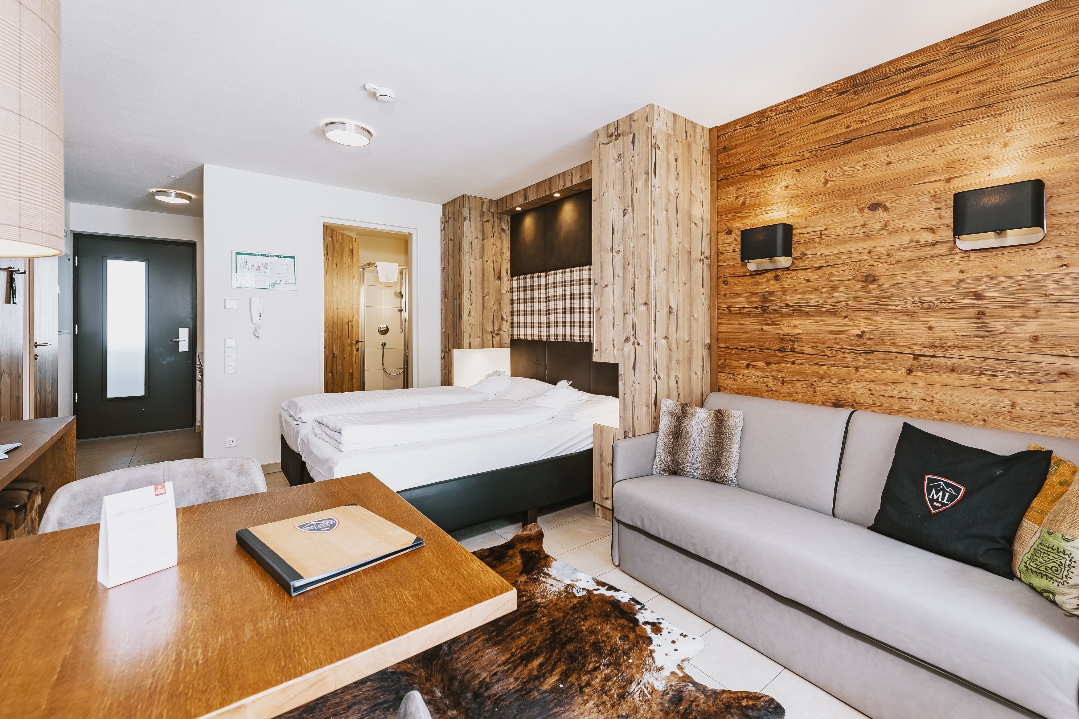 A modern hotel room with a wooden accent wall, a bed, a sofa, a desk, and a cowhide rug.