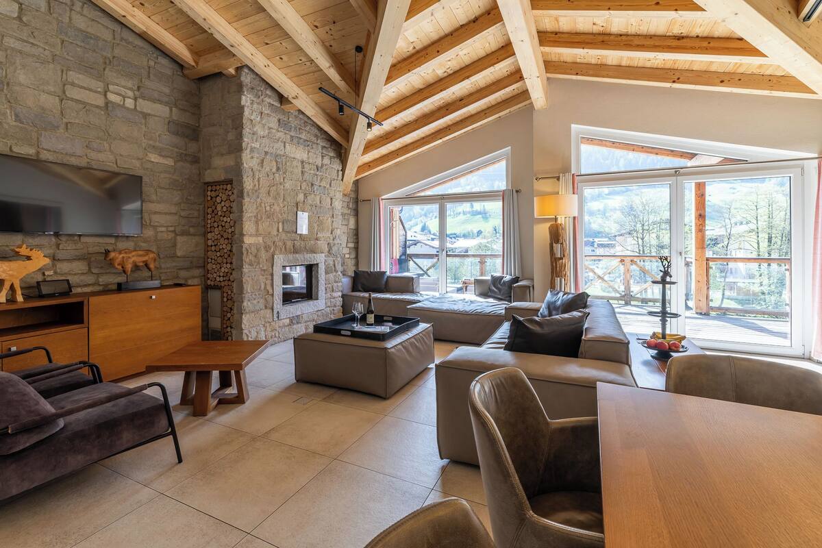 A spacious living area with a stone fireplace, wooden beams, a flat-screen TV, and large windows offering a view of the outdoors.