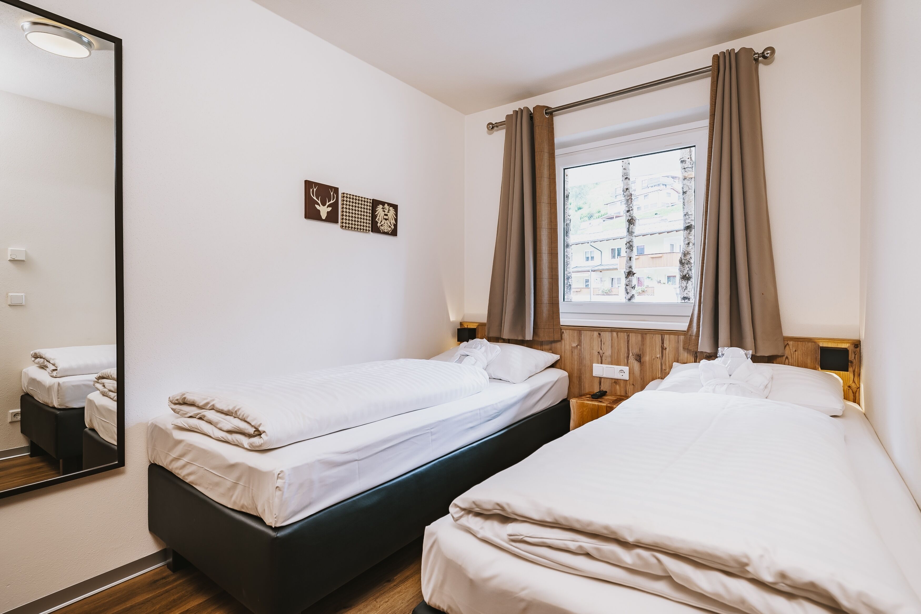 A hotel room with two single beds, a large mirror, a window with curtains, and a wall-mounted artwork.