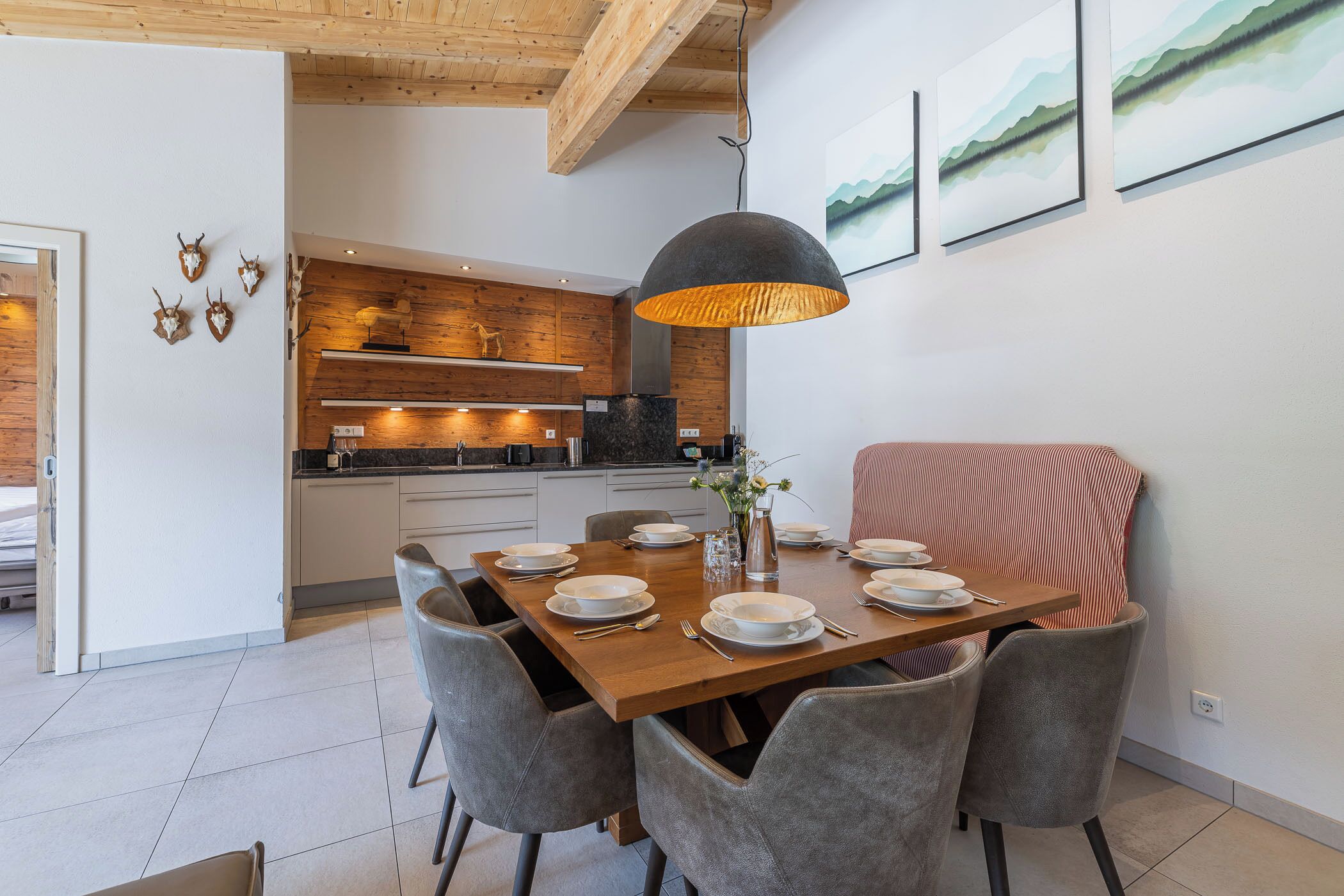 Un comedor con una mesa de madera dispuesta para cuatro personas, sillas grises tapizadas, una lámpara colgante y una cocina al fondo.