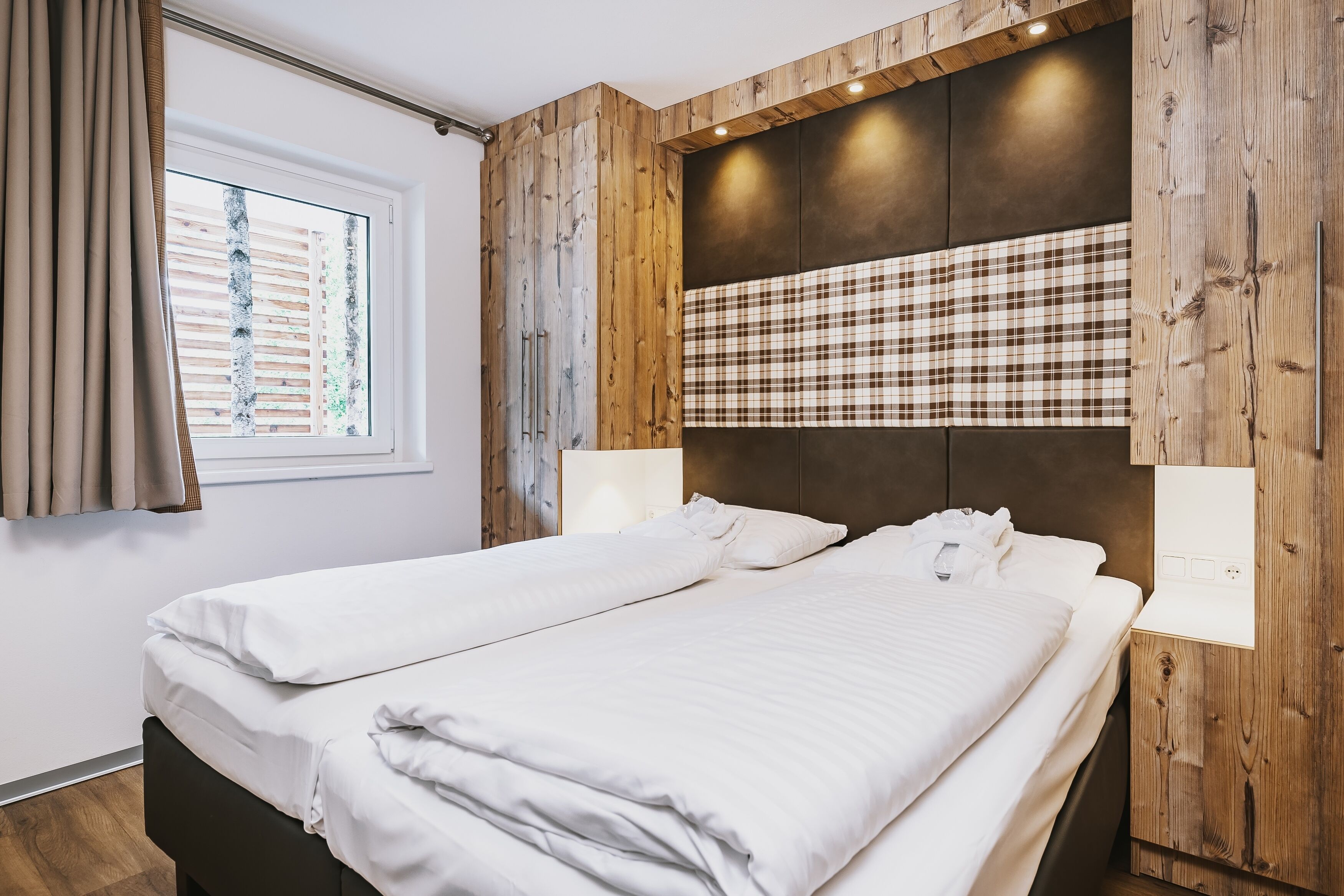 Een tweepersoonsbed met witte lakens, een raam met zonwering en een houten hoofdbord met een geruit paneel.