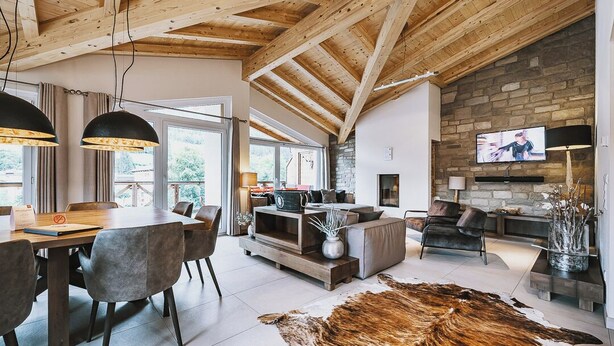 Un vaste espace de vie doté d’un plafond en bois, d’une cheminée en pierre, d’un téléviseur et d’un coin repas avec une table et des chaises.