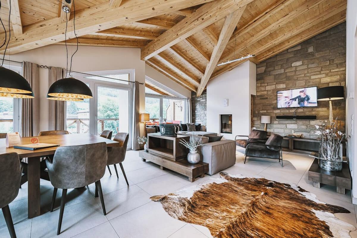 A spacious living area with a wooden ceiling, a stone fireplace, a television, and a dining area with a table and chairs.