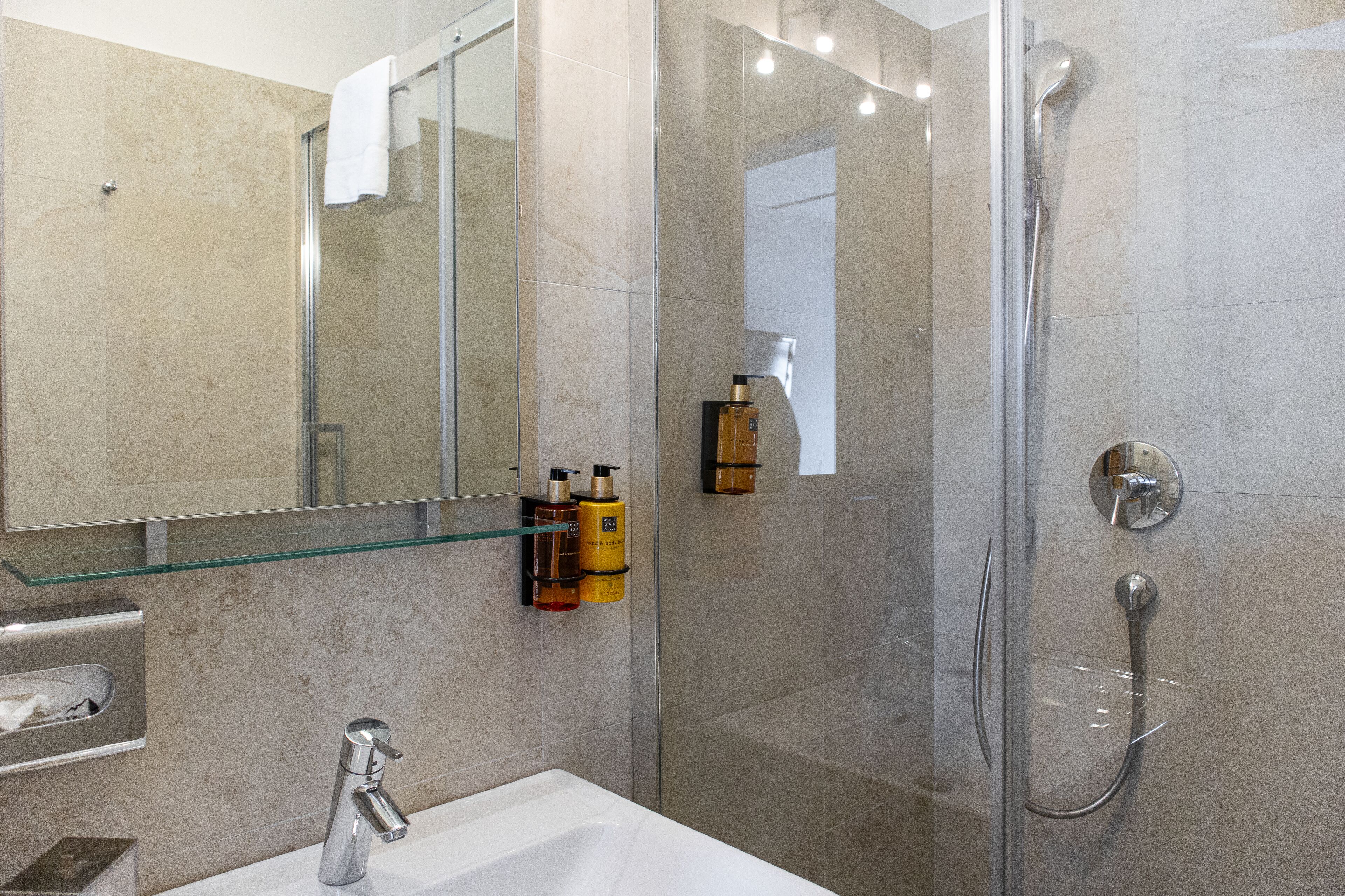 A modern bathroom with a glass shower enclosure, a white sink, and a glass shelf with toiletries.