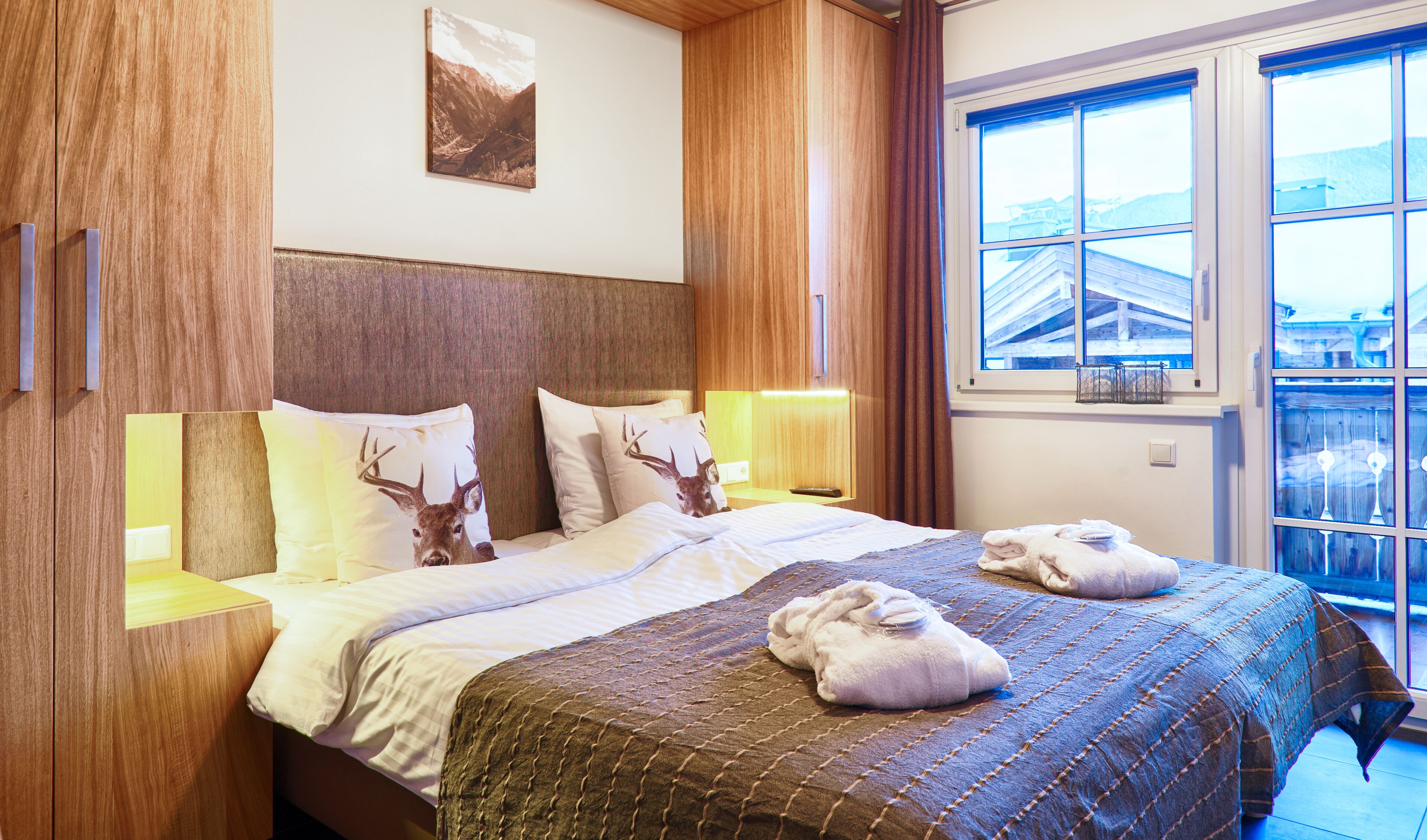 Une chambre d’hôtel avec un grand lit, une tête de lit en bois, deux oreillers ornés de motifs de cerfs, une couverture et une vue sur un immeuble par la fenêtre.