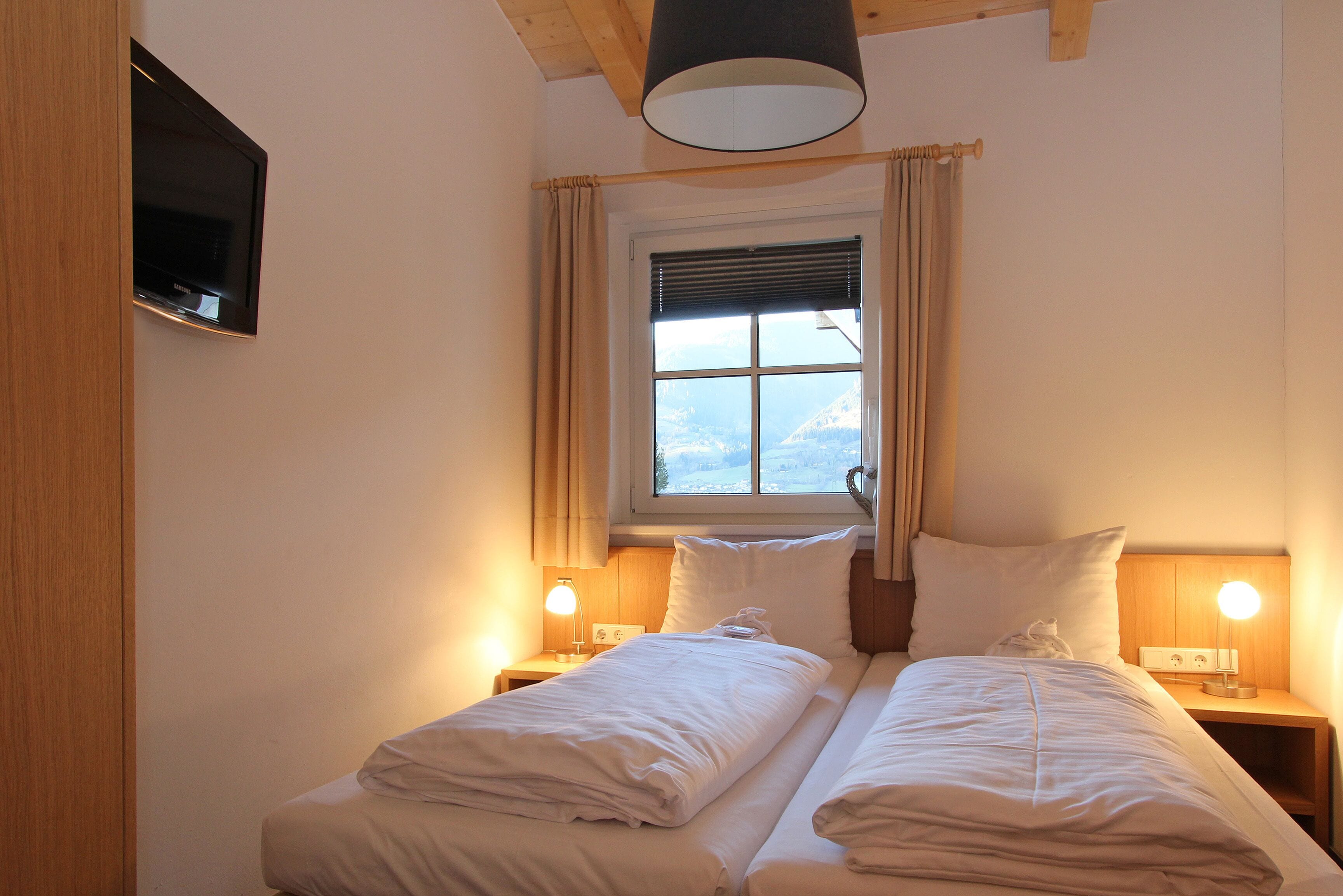 A bedroom with two beds, a TV mounted on the wall, a window with curtains, and bedside lamps.