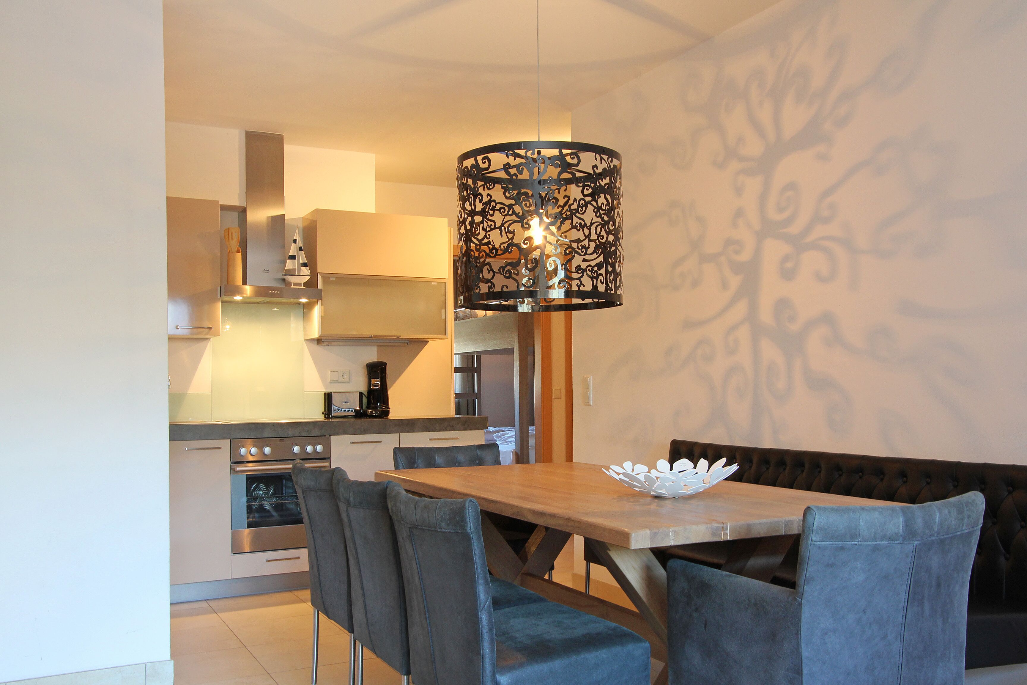 A modern kitchen with stainless steel appliances, a dining area with a wooden table and blue chairs, and a decorative wall with a tree pattern.