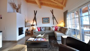 A living room with a wooden ceiling, a fireplace, a sofa, and a large window. - AvenidA Mountain Resort by Alpin Rentals - incl Summercard (Kaprun)