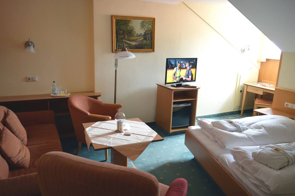 Hotellihuone, jossa on sänky, työpöytä tietokoneella, tuoli, pöytä pullolla ja laseilla sekä taulu seinällä.