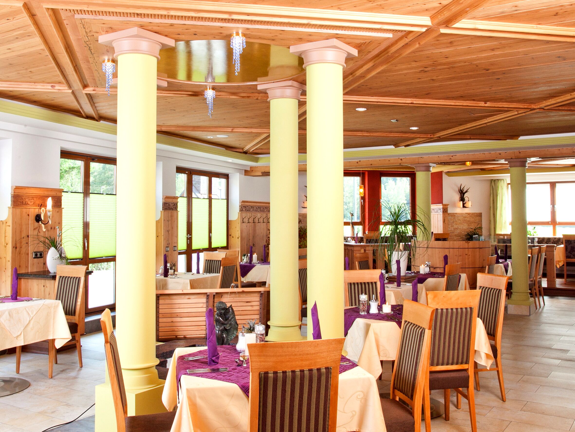 Un restaurante con vigas y columnas de madera, mesas con servilletas moradas y vistas al exterior a través de amplios ventanales.