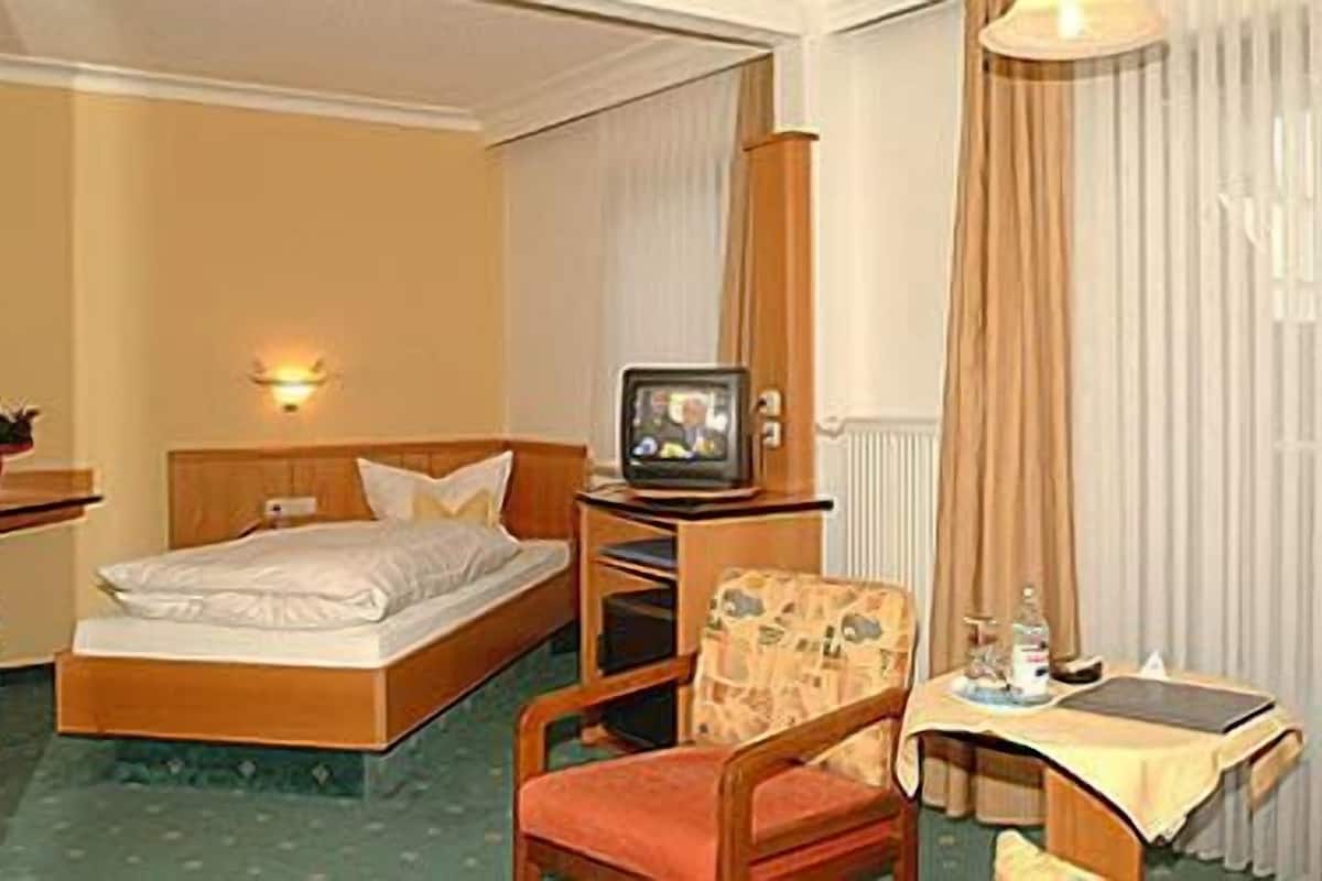 Hotellihuone, jossa on sänky, puinen jalusta, jonka päällä on televisio, tuoli ja pieni pöytä, jossa on lamppu.