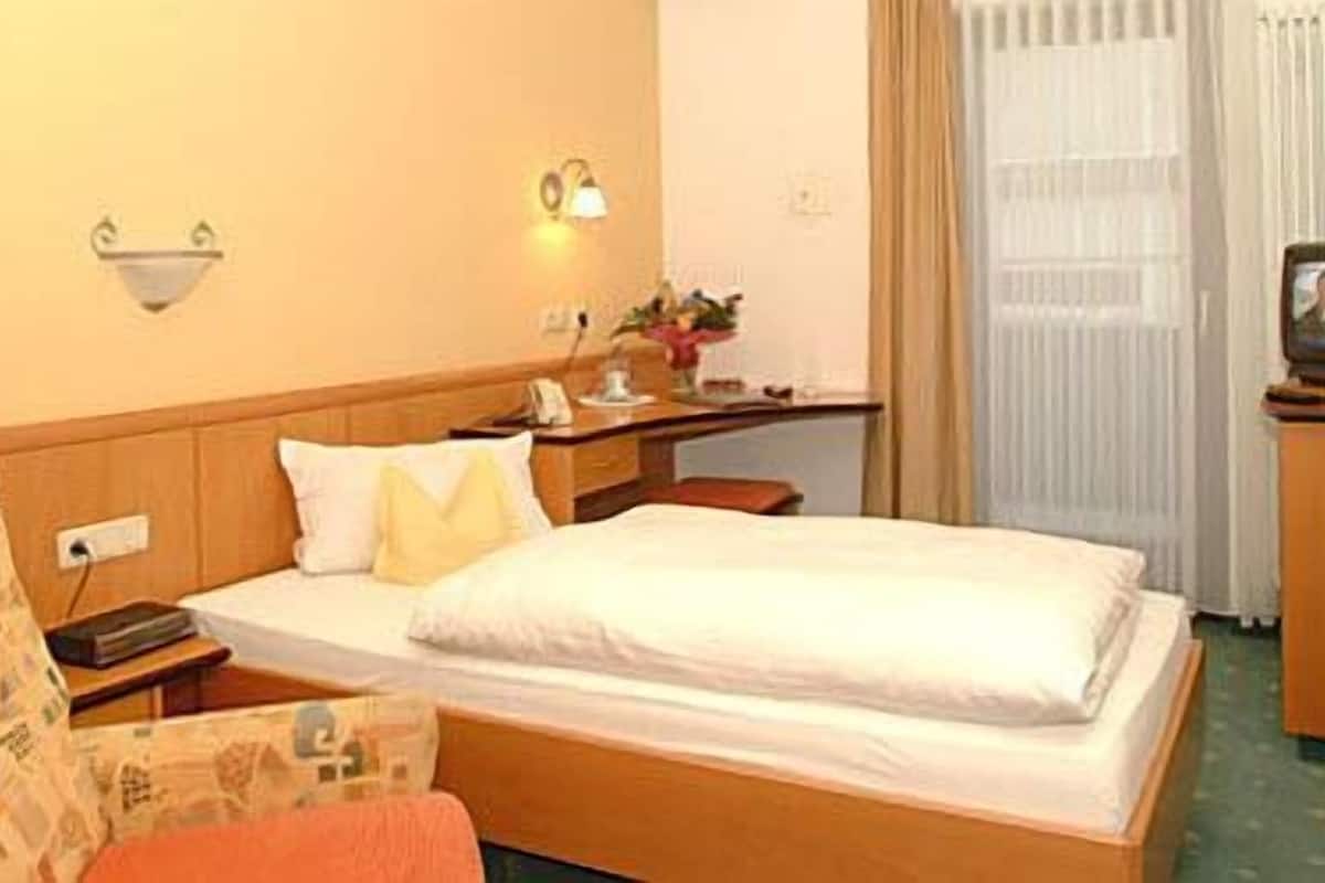 Hotellihuone, jossa on yksi sänky, yöpöytä, tuoli, työpöytä, lamppu, televisio ja ikkuna, jossa on verhot.