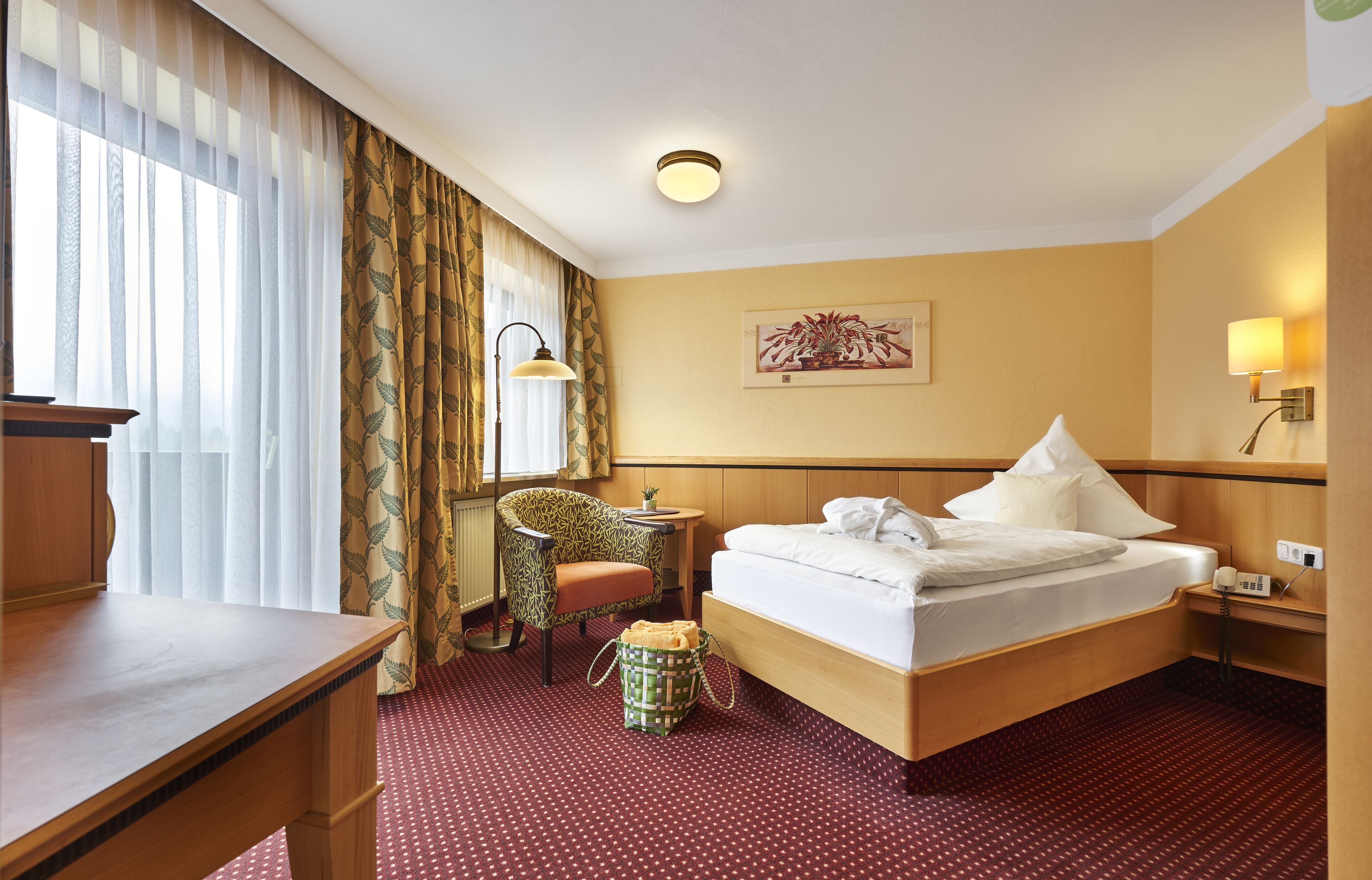 A hotel room with a bed, a desk, a chair, a small table, a lamp, and a painting on the wall.