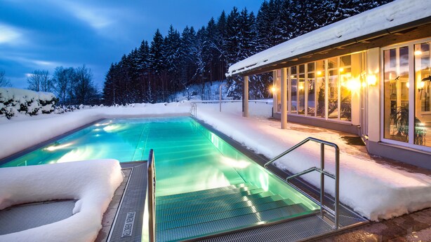 Una piscina con cerramiento de vidrio, rodeada de suelo y árboles cubiertos de nieve, y un edificio con amplias ventanas y luces en el fondo.