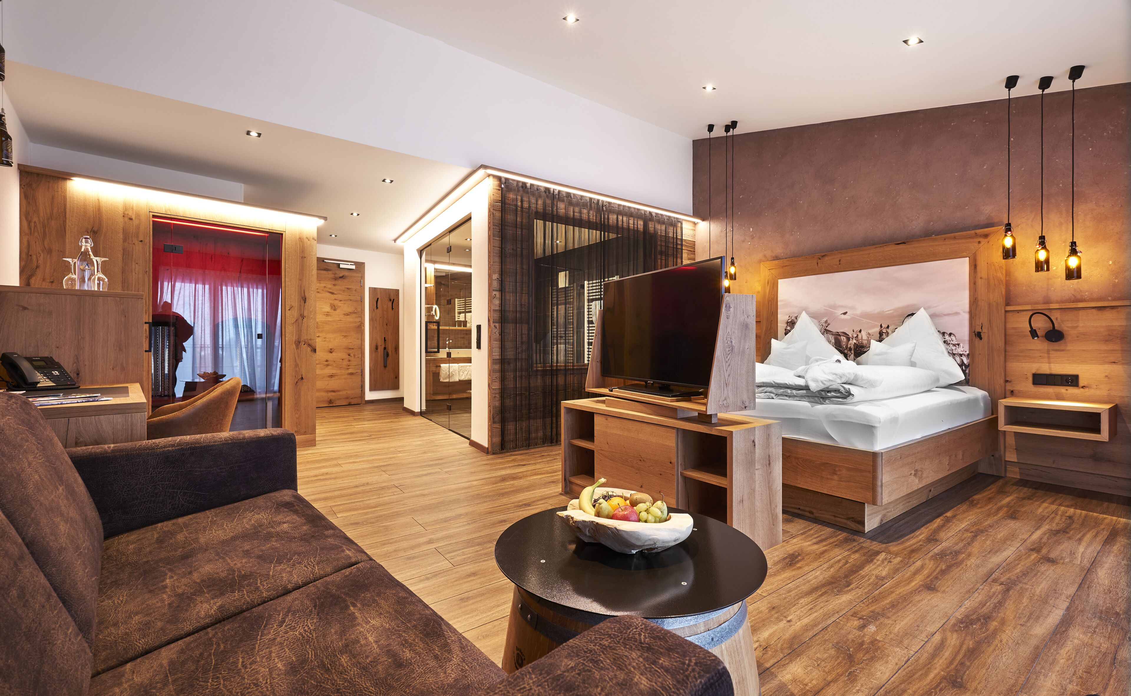 A modern hotel room with a wooden floor, a large bed, a TV, a desk, and a sofa.