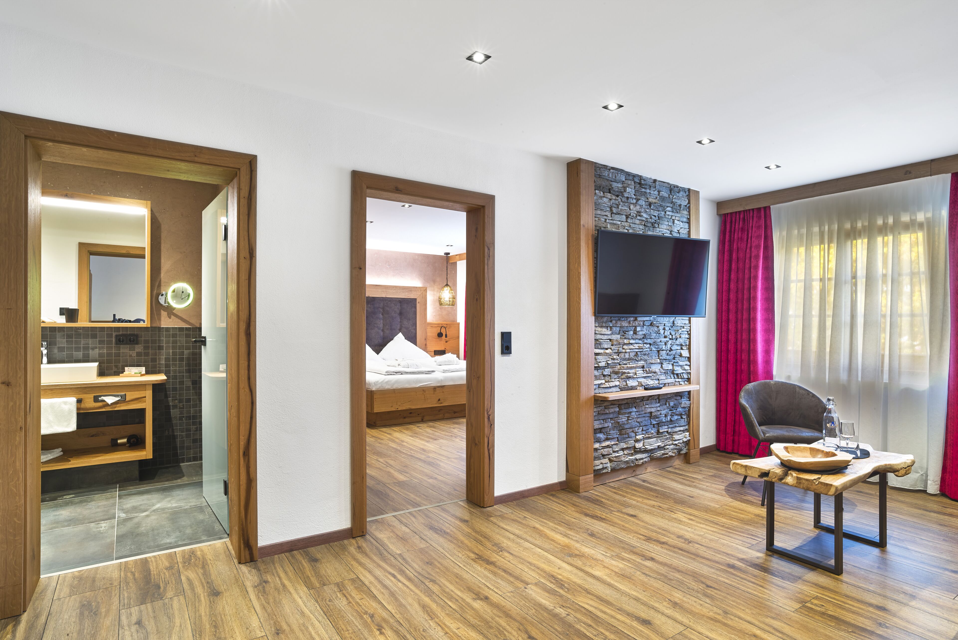 Ein modernes Hotelzimmer mit Holzboden, einer Wand mit Steinakzenten, einem Flachbildfernseher, einem gemütlichen Bett, einer Holzbank mit Schale und einem großen Fenster mit Vorhängen.
