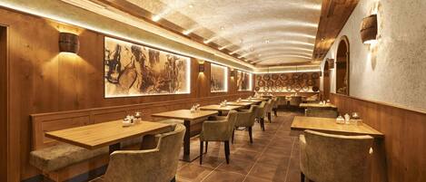 Un restaurant avec des tables et des chaises en bois, un plafond haut avec des poutres apparentes et des luminaires fixés aux murs.