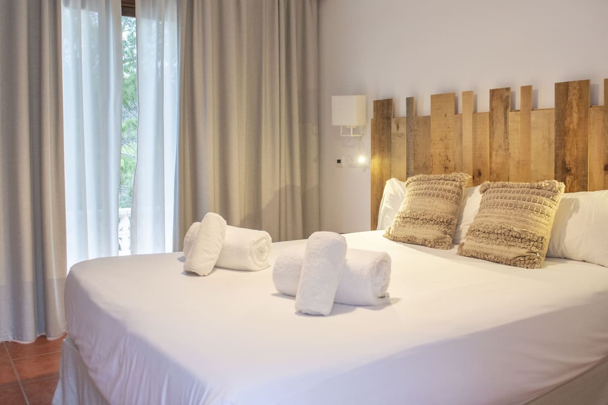 Een opgemaakt bed met witte lakens en opgerolde handdoeken, een houten hoofdbord en een raam met dunne gordijnen.