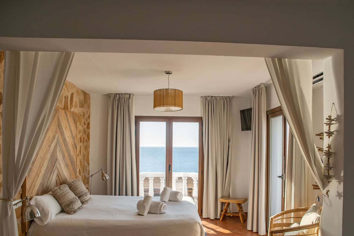 Een slaapkamer met een groot bed, een houten hoofdbord, een bedlampje, een klein houten krukje en uitzicht op de zee door een glazen deur.