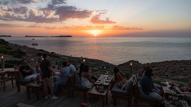 Vista del atardecer desde una terraza con mesas y sillas, con vista al mar.