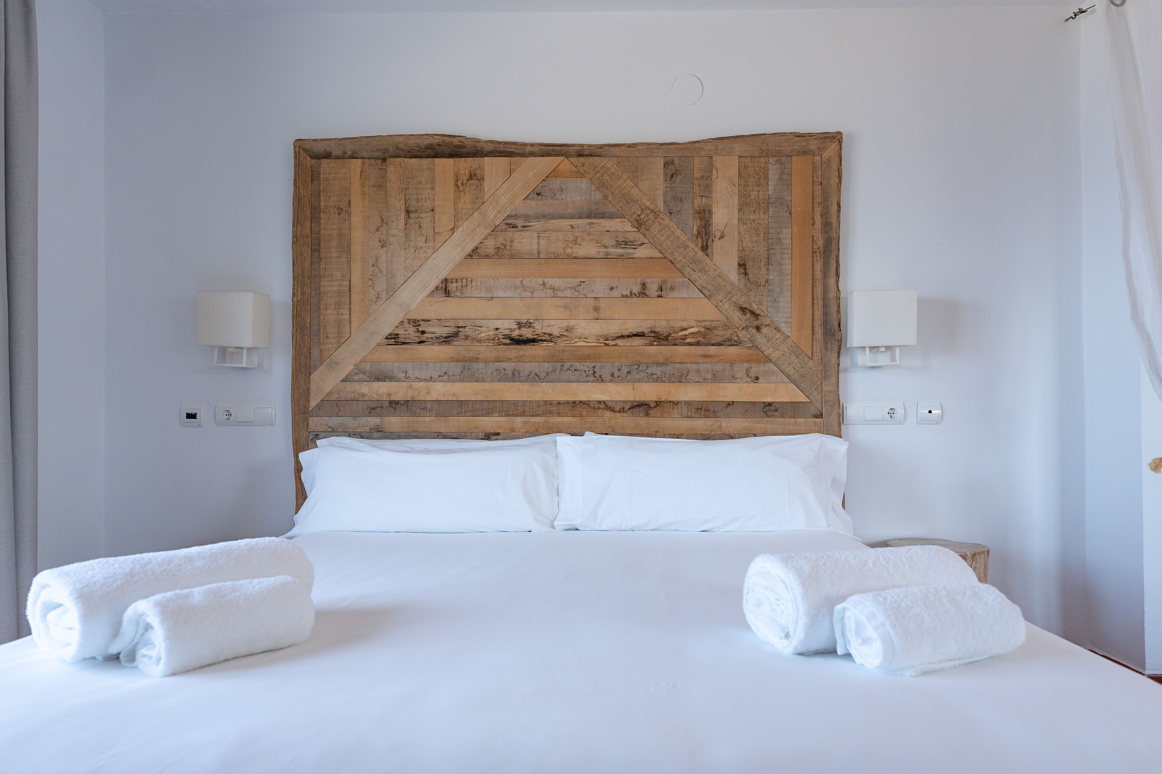 A bedroom with a wooden headboard, white bedding, two bedside lamps, and two rolled towels on the bed.