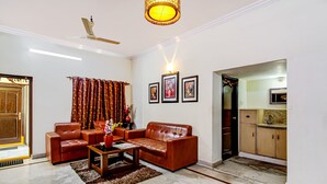 A living room with a brown leather sofa, a coffee table, and a ceiling fan. There are framed pictures on the wall and a door leading to a kitchen.