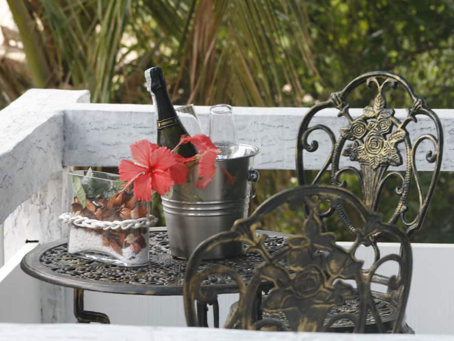 Un balcón con una silla metálica, barandilla decorativa de metal y una pequeña mesa con una botella y flores.