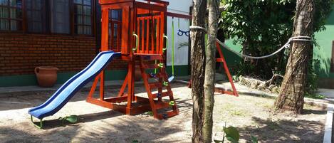 Un parque infantil con tobogán y estructura para escalar, rodeado de árboles y un edificio al fondo.