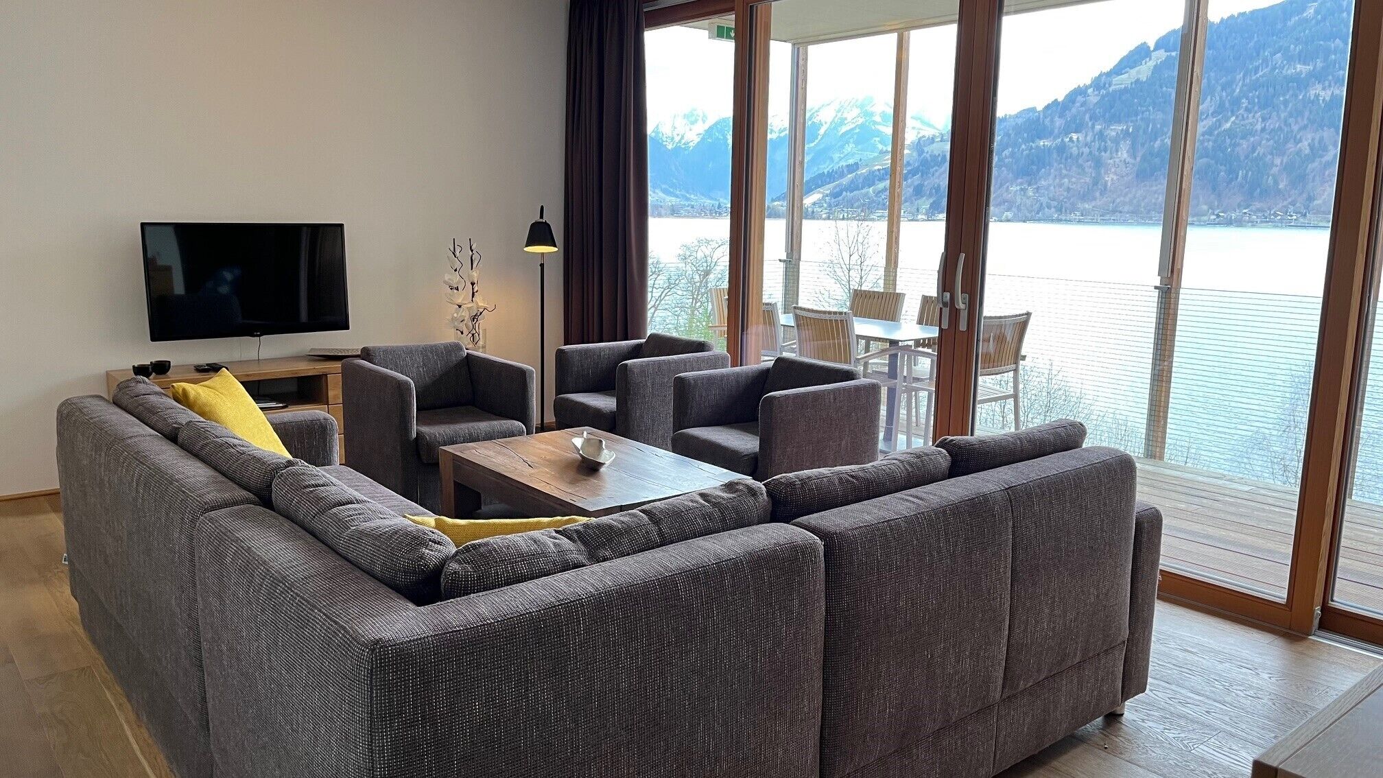 Una sala moderna con un sofá gris, una mesa de centro de madera y un televisor de pantalla plana. Ventanas amplias ofrecen vistas a un lago y montañas.