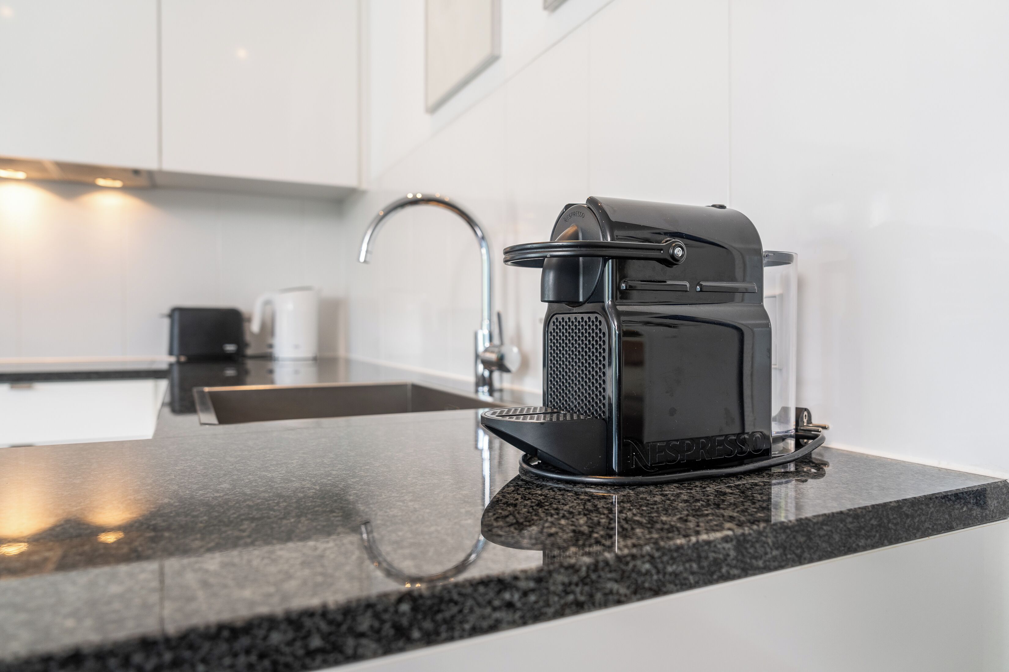 Una cucina moderna con una macchina da caffè nera su un piano di lavoro in granito, un rubinetto in acciaio inossidabile e mobili bianchi.