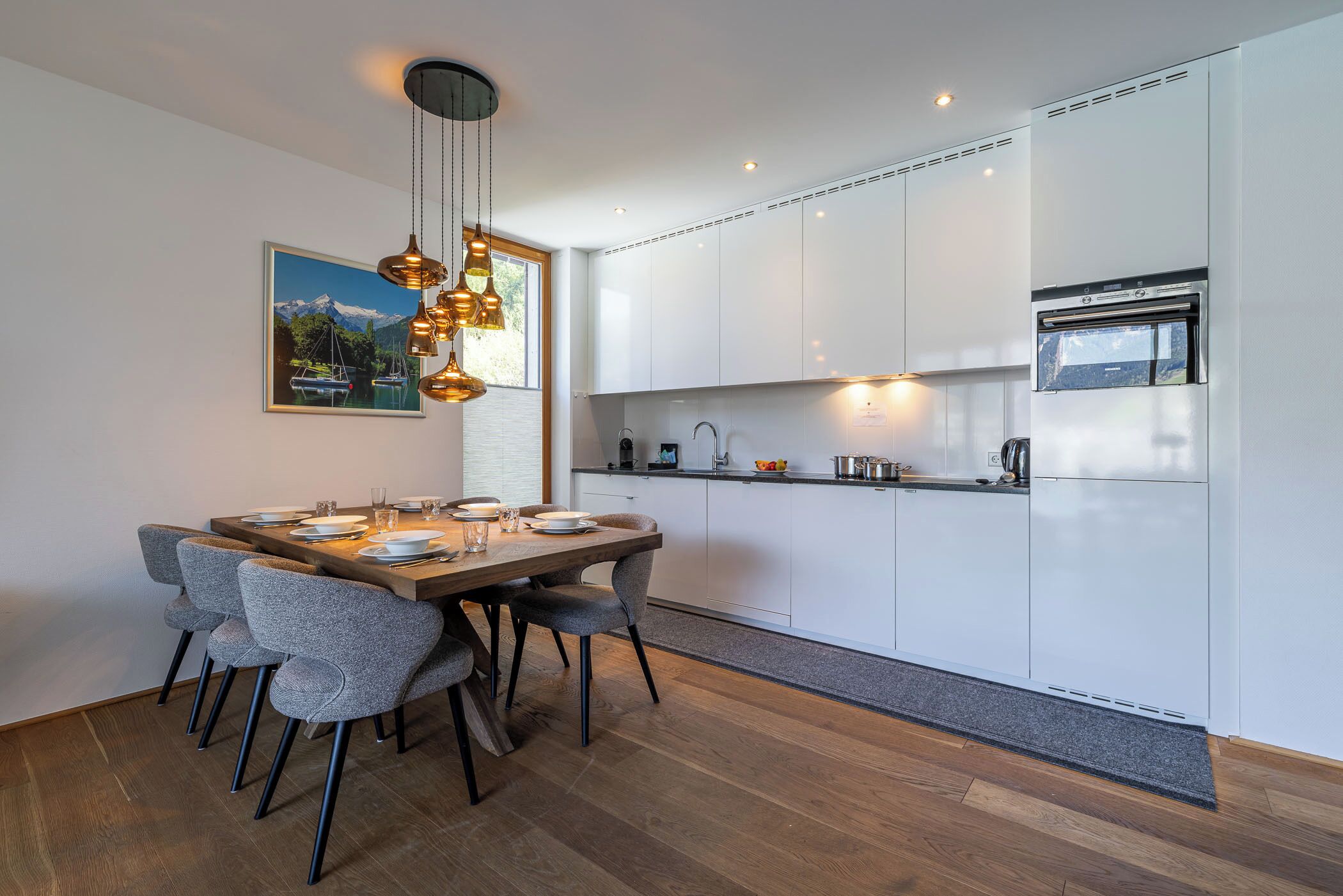 Cocina moderna con armarios blancos, horno empotrado y comedor con mesa de madera y sillas grises.