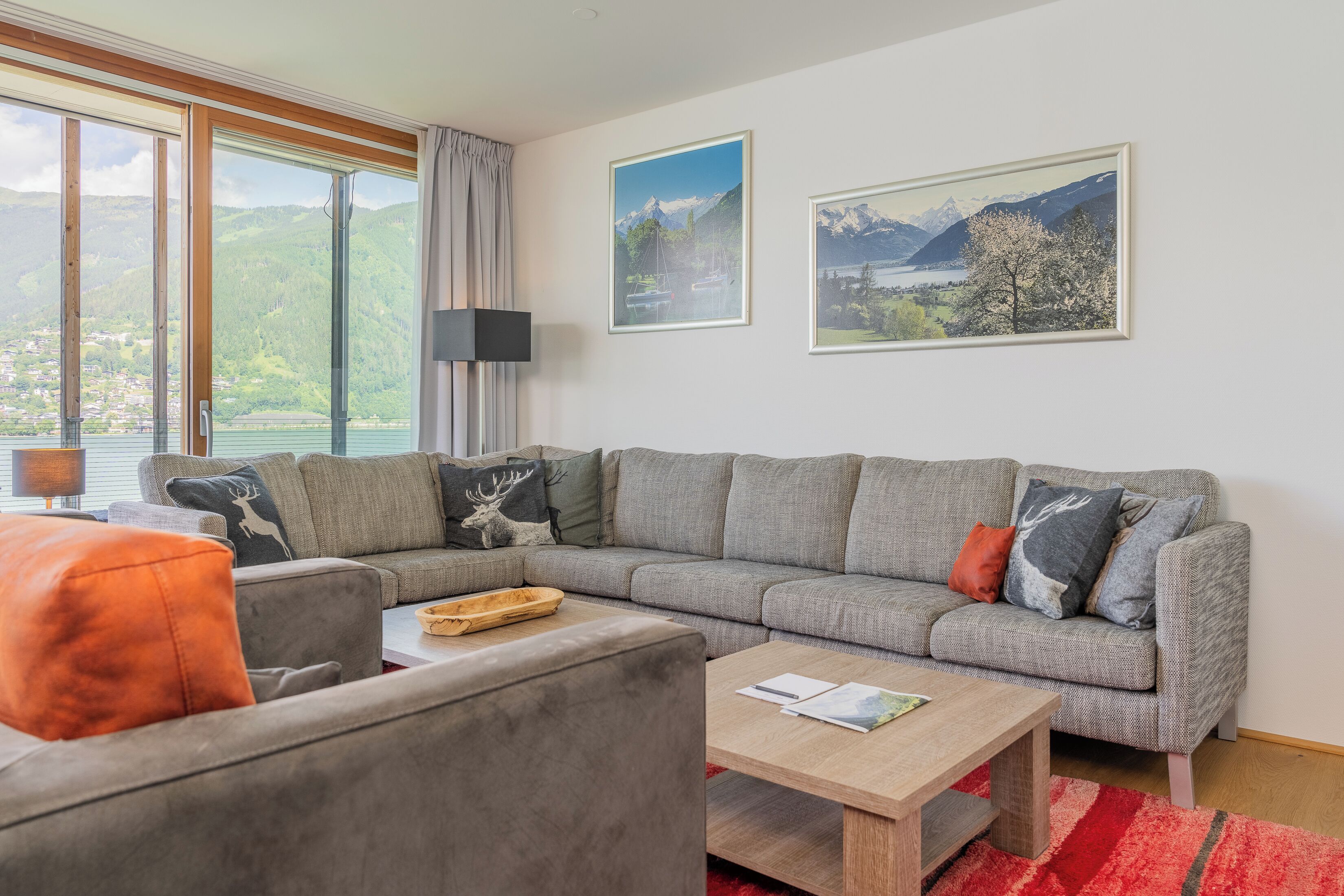 Una sala moderna con un sofá seccional gris, una mesa de centro de madera y una alfombra roja. Hay dos cuadros con paisajes enmarcados en la pared y una ventana con vistas a montañas y un lago.