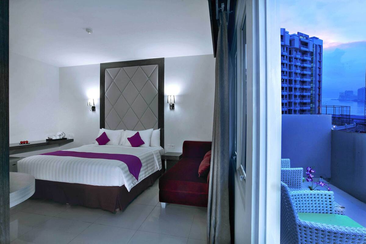 A hotel room with a bed, a red chair, and a view of a cityscape.