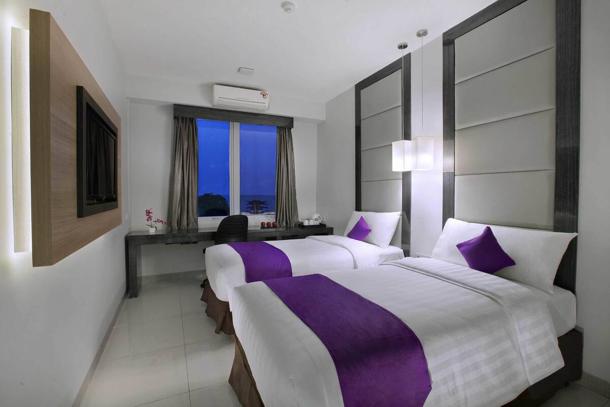 A hotel room with two beds, a desk, a chair, a window with curtains, and an air conditioning unit.