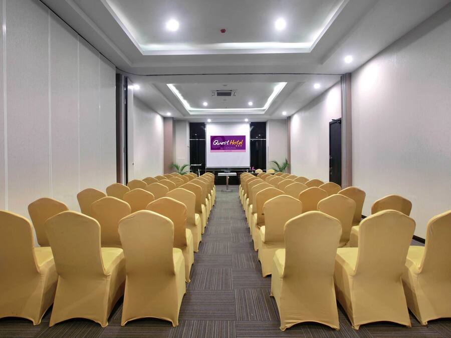 Une salle de conférence avec des rangées de chaises jaunes, un pupitre et un écran affichant le nom « Quest Hotel ».