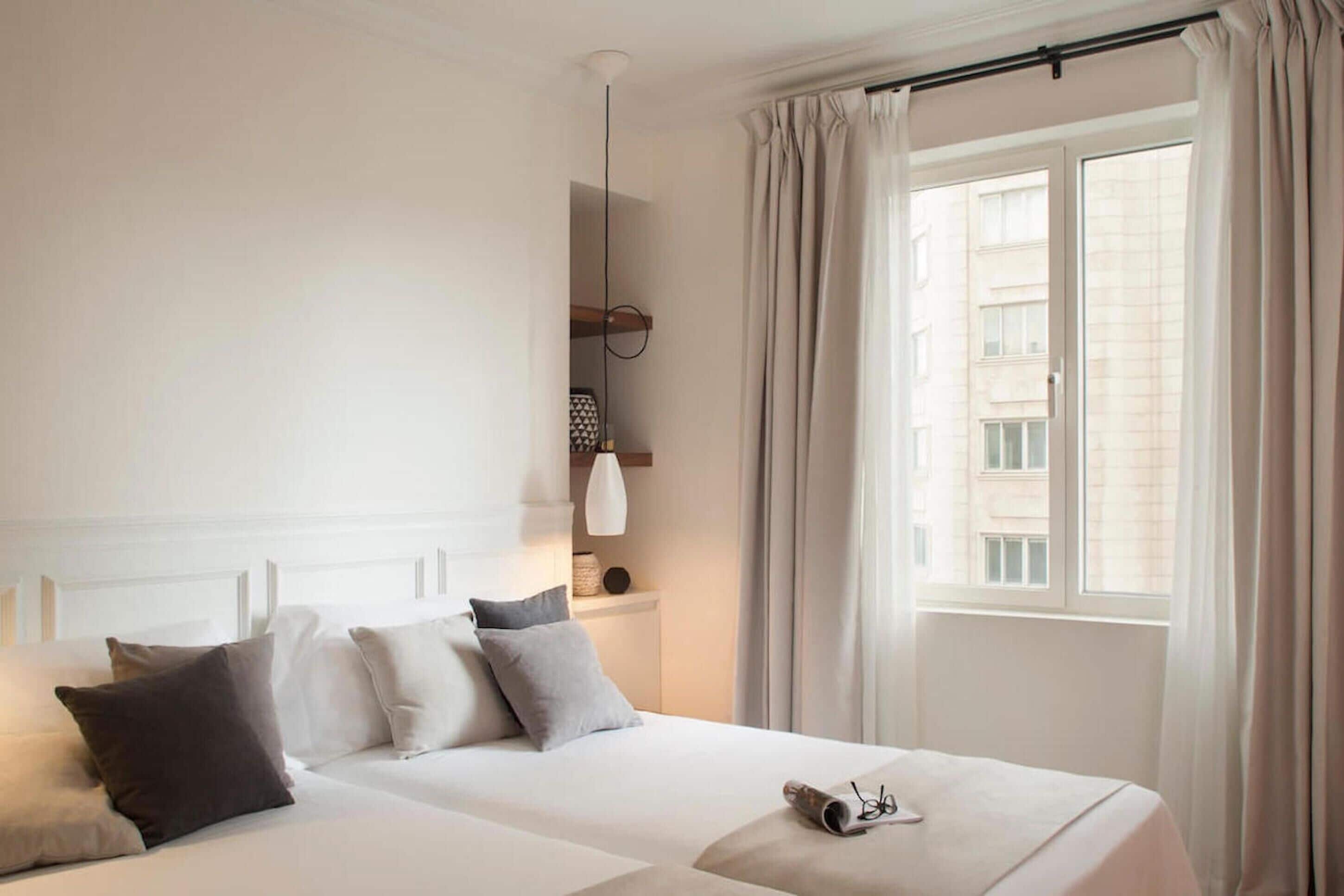 Habitación de hotel con dos camas, una mesita de noche con lámpara, una ventana con cortinas y vistas a edificios.
