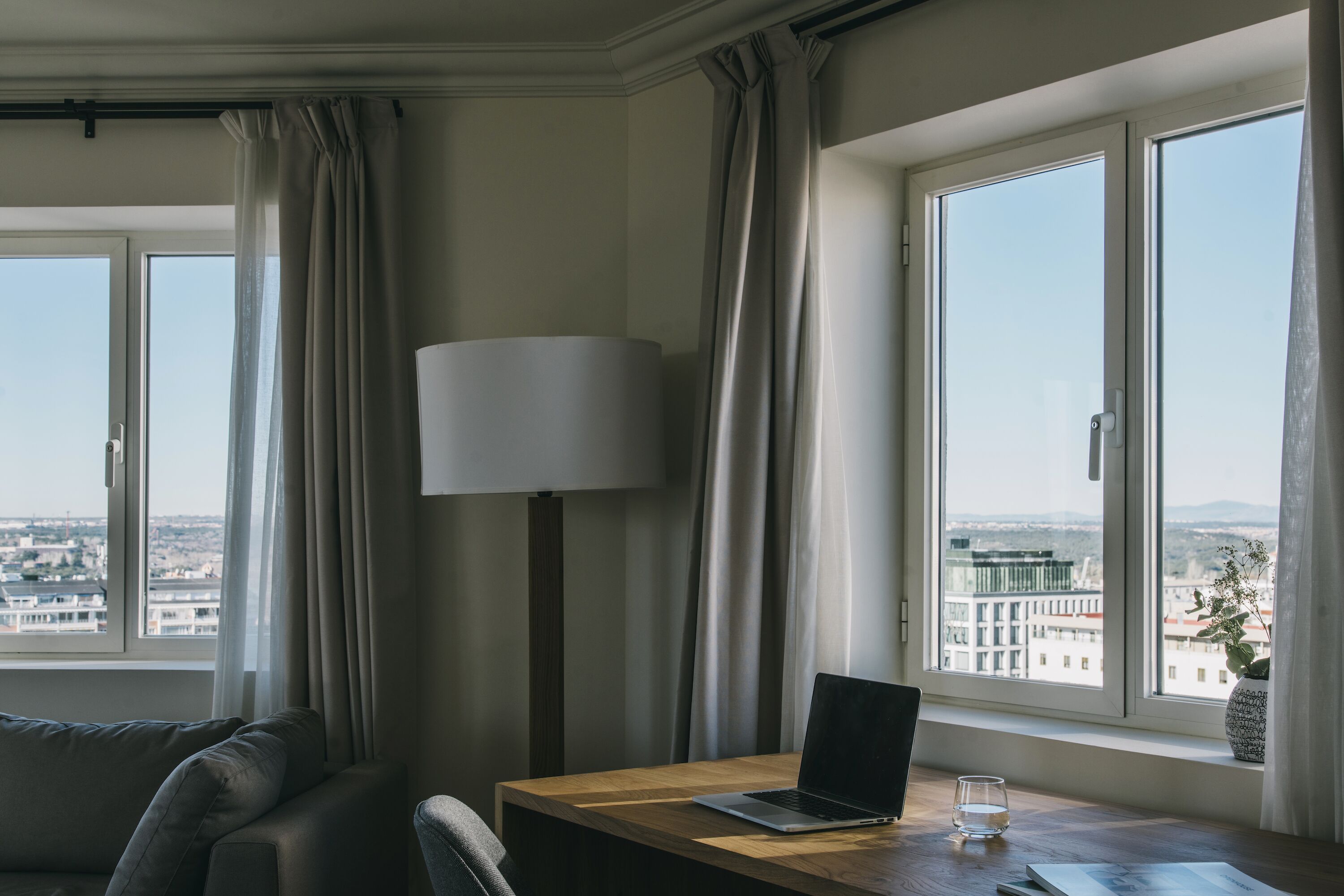 Een moderne hotelkamer met een groot raam, een houten bureau, een laptop, een lamp en uitzicht op de stad.