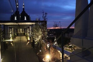 A rooftop terrace with potted plants, a covered seating area, and a view of a cityscape at dusk. - Dear Hotel Madrid (Madrid)
