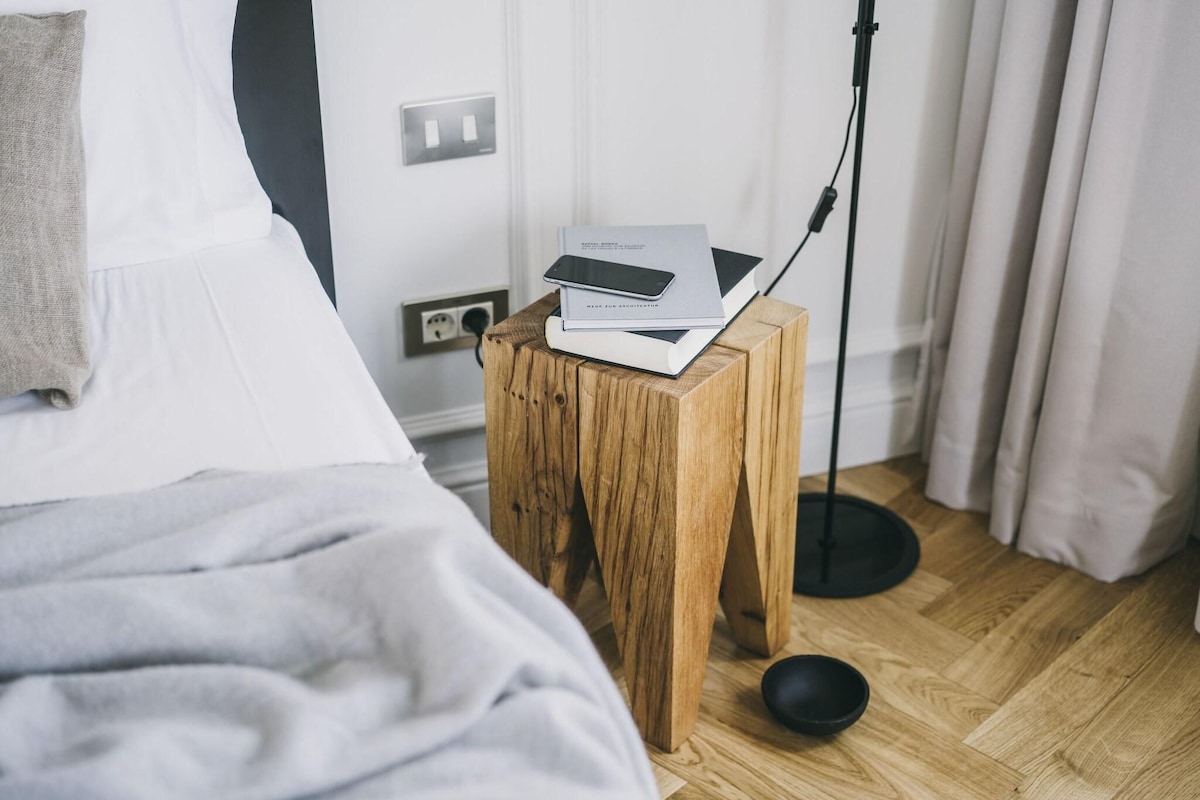 A neatly made bed with a gray blanket, a wooden bedside table with a smartphone and a book, a floor lamp, and a power outlet on a wooden floor.