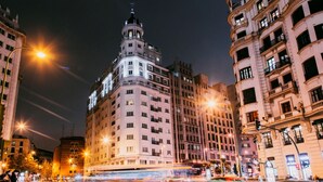 A nighttime cityscape with illuminated buildings, a prominent tower with a rounded top, and a busy street with moving vehicles. - Dear Hotel Madrid (Madrid)