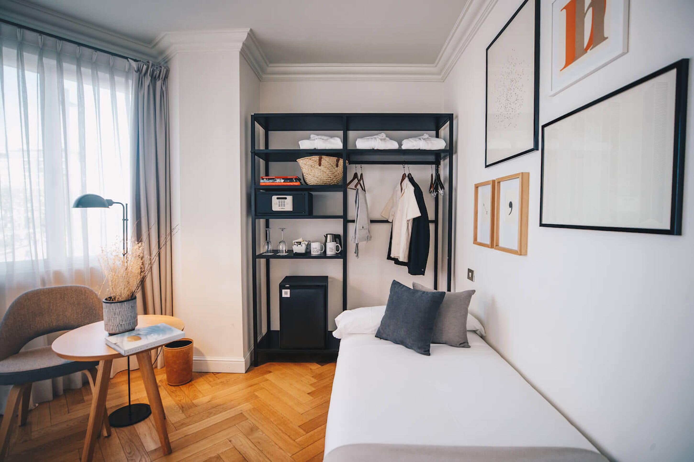 Een knus hotelkamer met een bed, een klein tafeltje met een lamp, een stoel, een kledingkast met hangende kleding en een minibar.